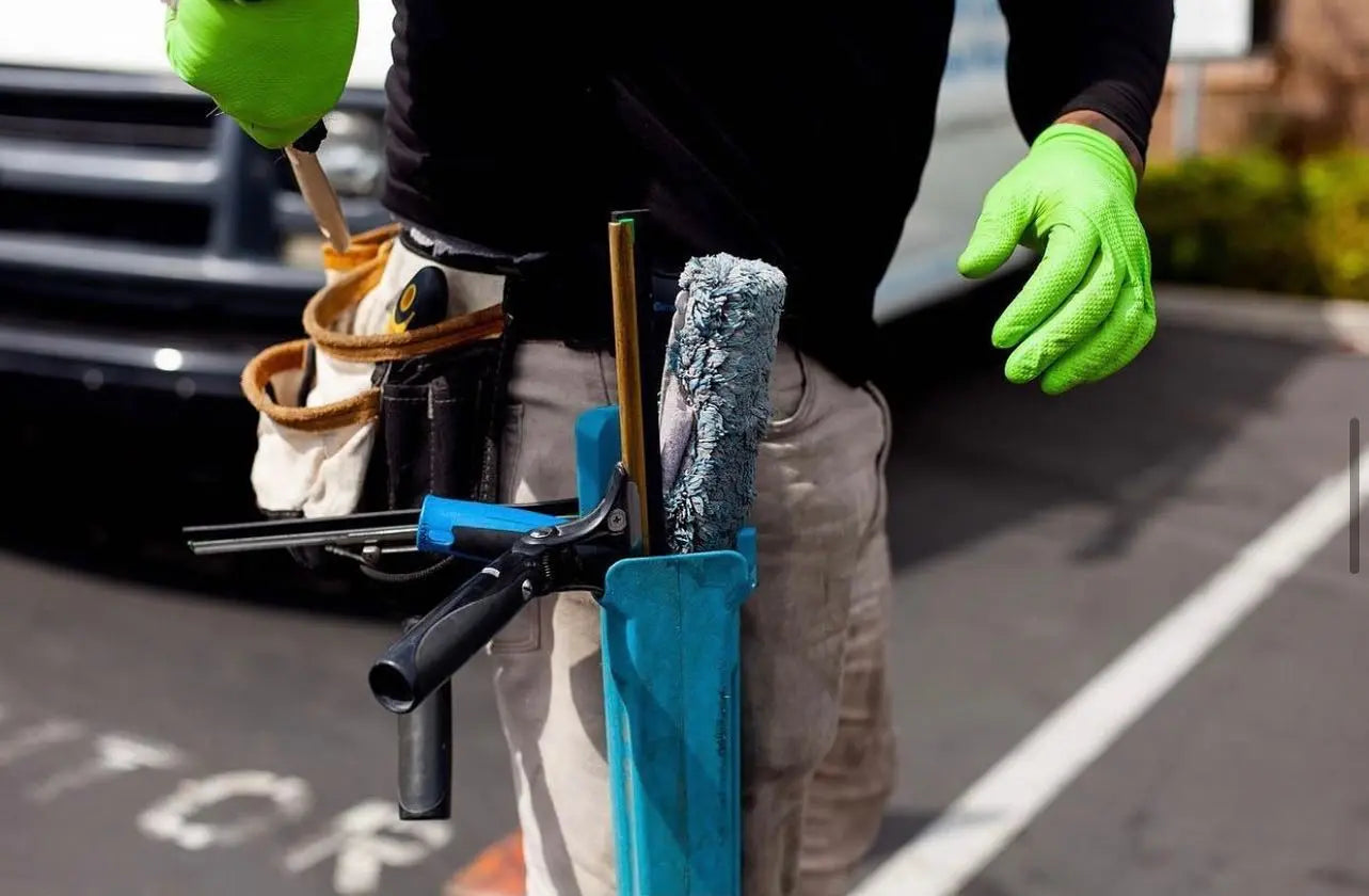 Person holding a tool belt with various tools, wearing green gloves, on a street.